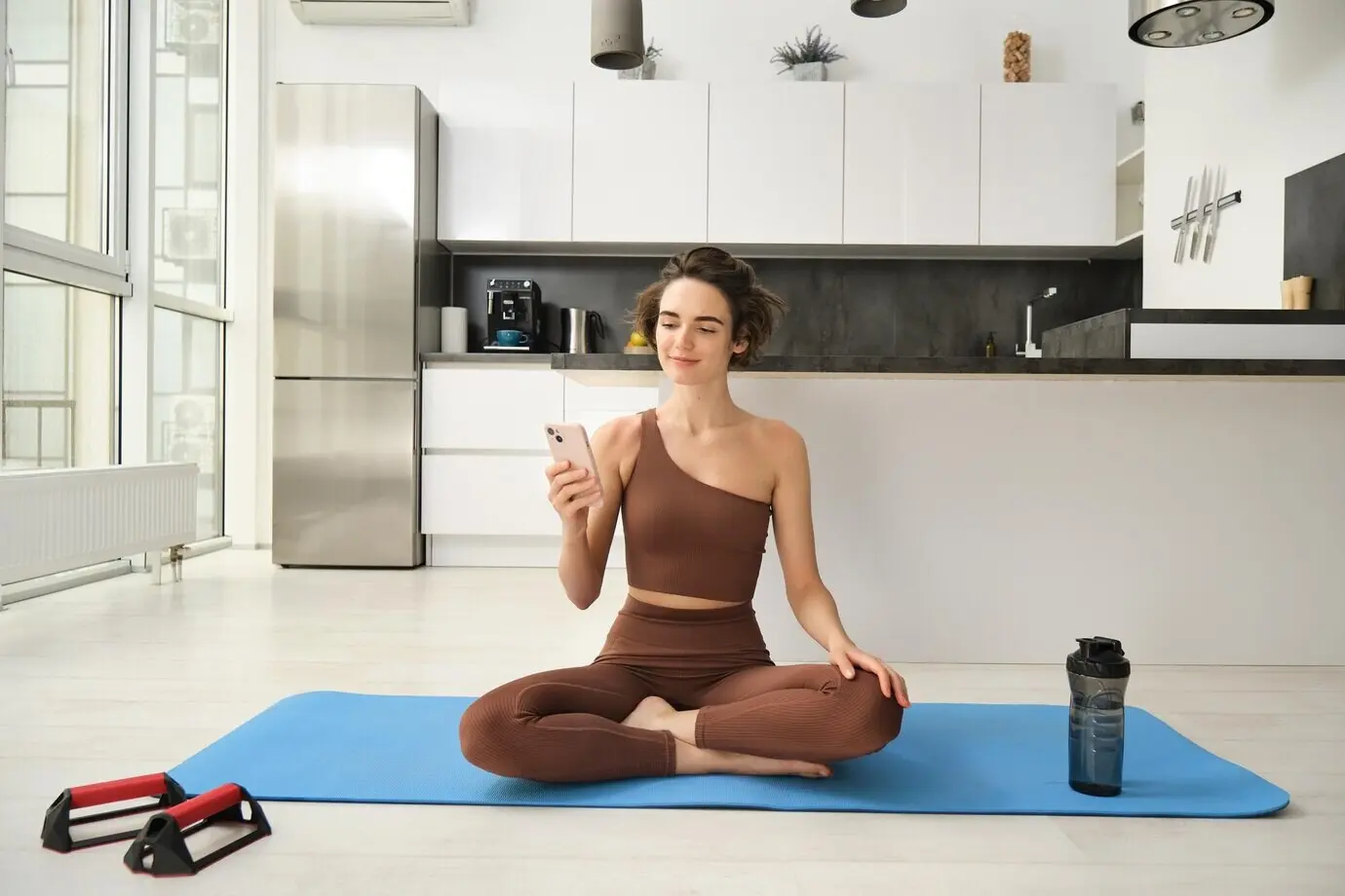 Sport-App und Wohlbefinden: Eine junge Sportlerin sitzt zu Hause auf dem Boden mit dem Smartphone und macht Yoga.